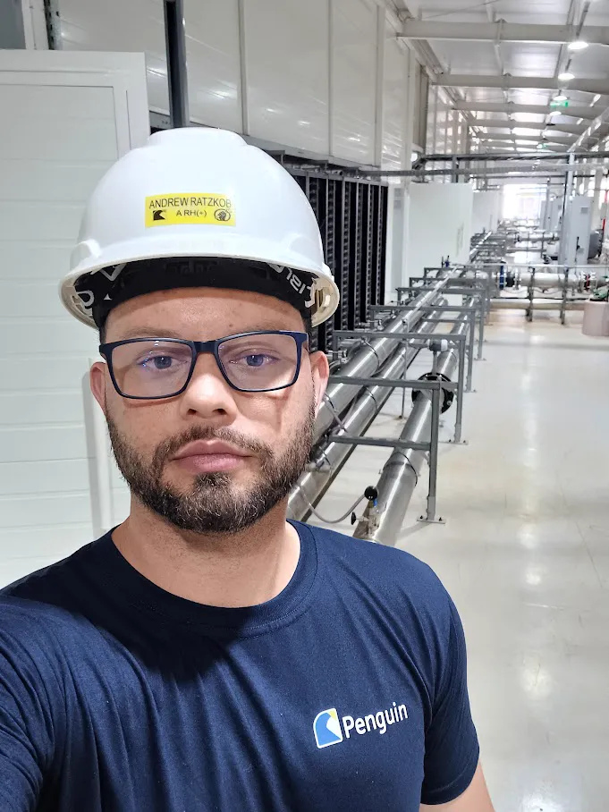 Andrew Ratzkob wearing a hard hat on site at a crypto/AI data center, next to rows of mining racks